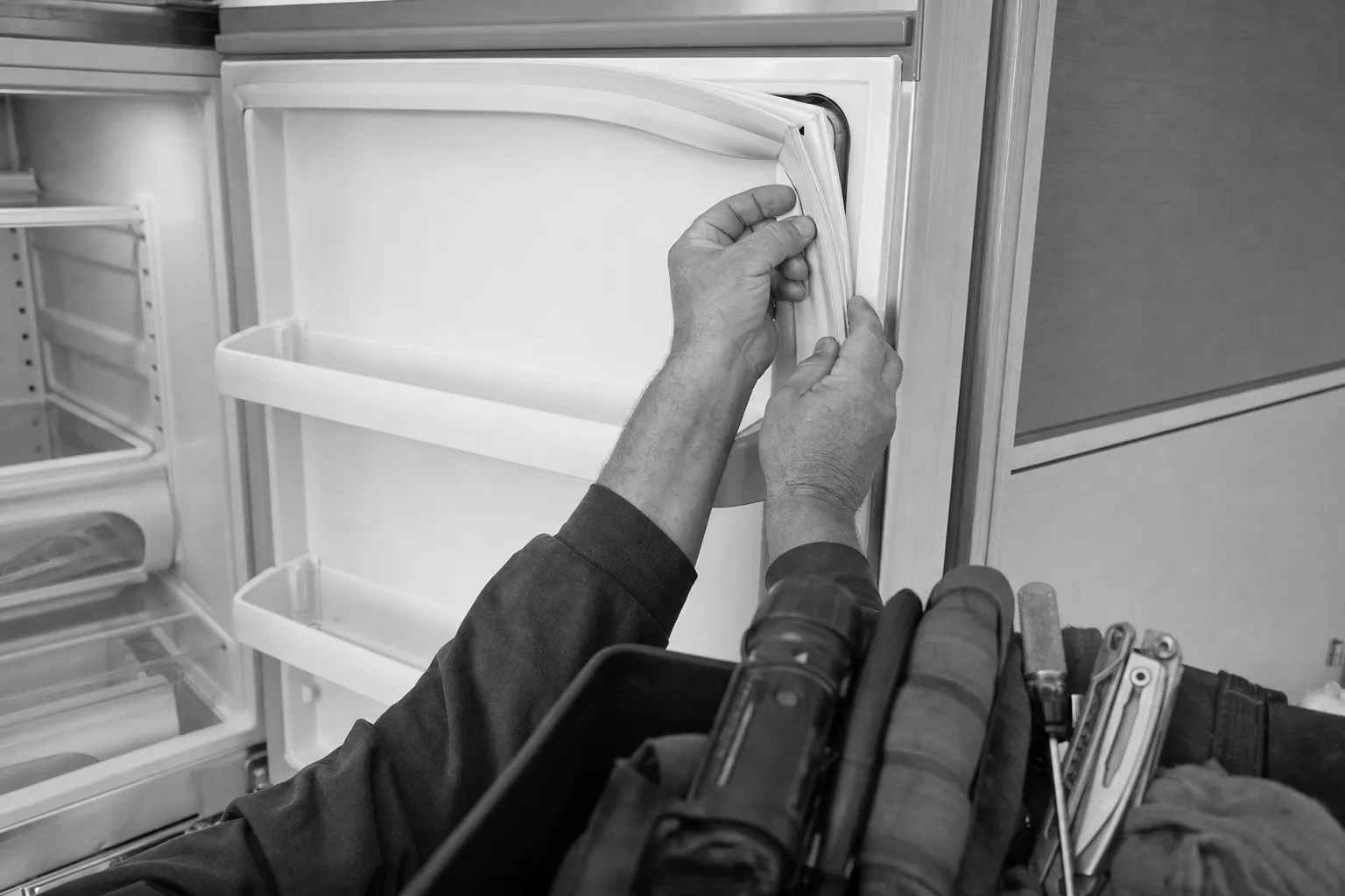 Black-and-white frame of both hands seating the gasket into the top corner of an open refrigerator door, a tool bag in front